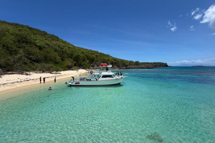 Boat anchored near a sandy beach with clear blue waters and green hills under a blue sky.