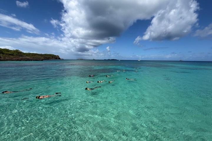 People snorkeling in clear turquoise water under a partly cloudy blue sky.