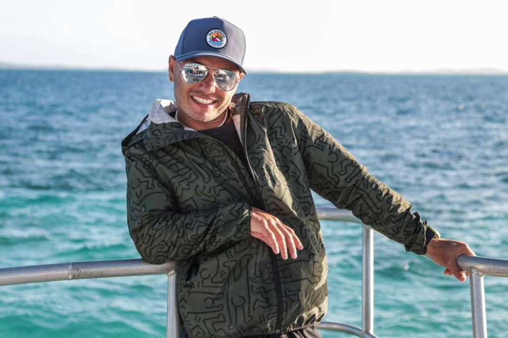 Smiling person in a jacket and cap standing by the ocean on a sunny day.